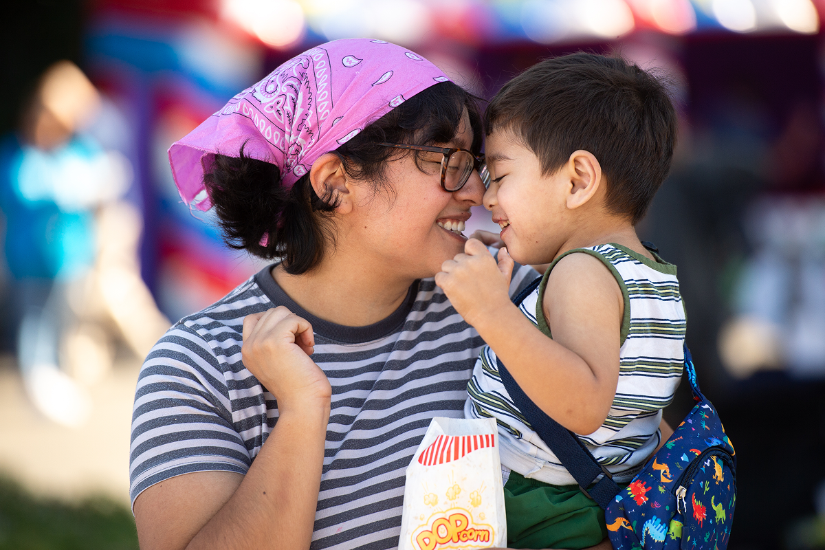 A woman in a striped shirt and pink bandana smiles as she touches noses with a young boy holding a popcorn bag at an outdoor event.