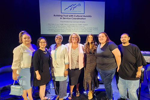 Seven people stand together on stage in front of a screen displaying a presentation titled 
