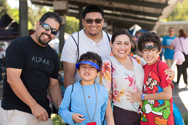 A group of five people, including two adults and three children, pose and smile outdoors at an event. One child is wearing face paint; another is holding cotton candy.