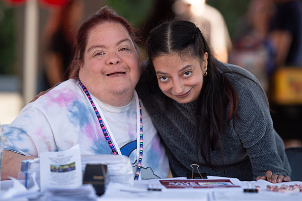 Two women smiling and posing together at a table covered with papers and brochures, outdoors in a casual setting.