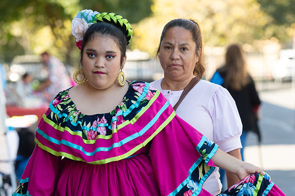 A young girl in a colorful traditional dress stands beside a woman in a light pink top outdoors during the day.